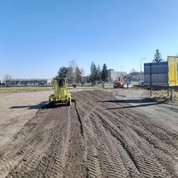 Firma Handlowo Usługowo Transportowa Krystian Bieniek - Żółty walec drogowy ugniata ziemię na placu budowy, widoczne ślady opon i oddalona koparka, w tle budynki i billboard reklamowy.