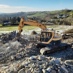 Firma Handlowo Usługowo Transportowa Krystian Bieniek - Żółta koparka Hyundai na gruzowisku po rozbiórce, w tle zielone wzgórza i fragmenty śniegu na łące.