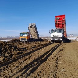 Firma Handlowo Usługowo Transportowa Krystian Bieniek - Dwa wywrotki z podniesionymi skrzyniami ładunkowymi na placu budowy, świeżo usypana ziemia z wyraźnymi śladami opon, błękitne niebo w tle.