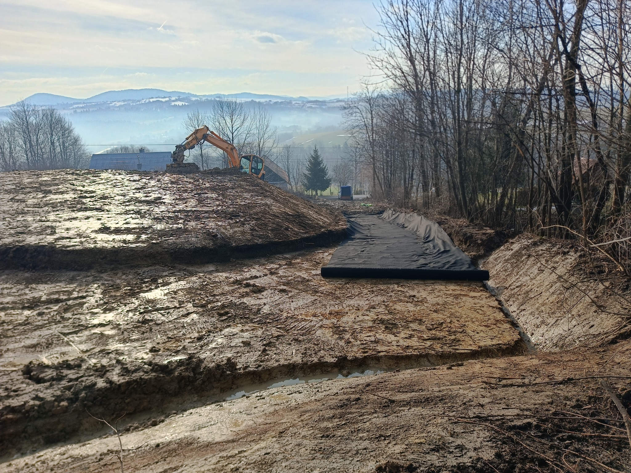 Żółta koparka w tle pracuje na błotnistym terenie; widoczne przygotowania pod warstwę drenażową w wykopie, z czarną geowłókniną; w oddali zalesione wzgórza pod błękitnym niebem.