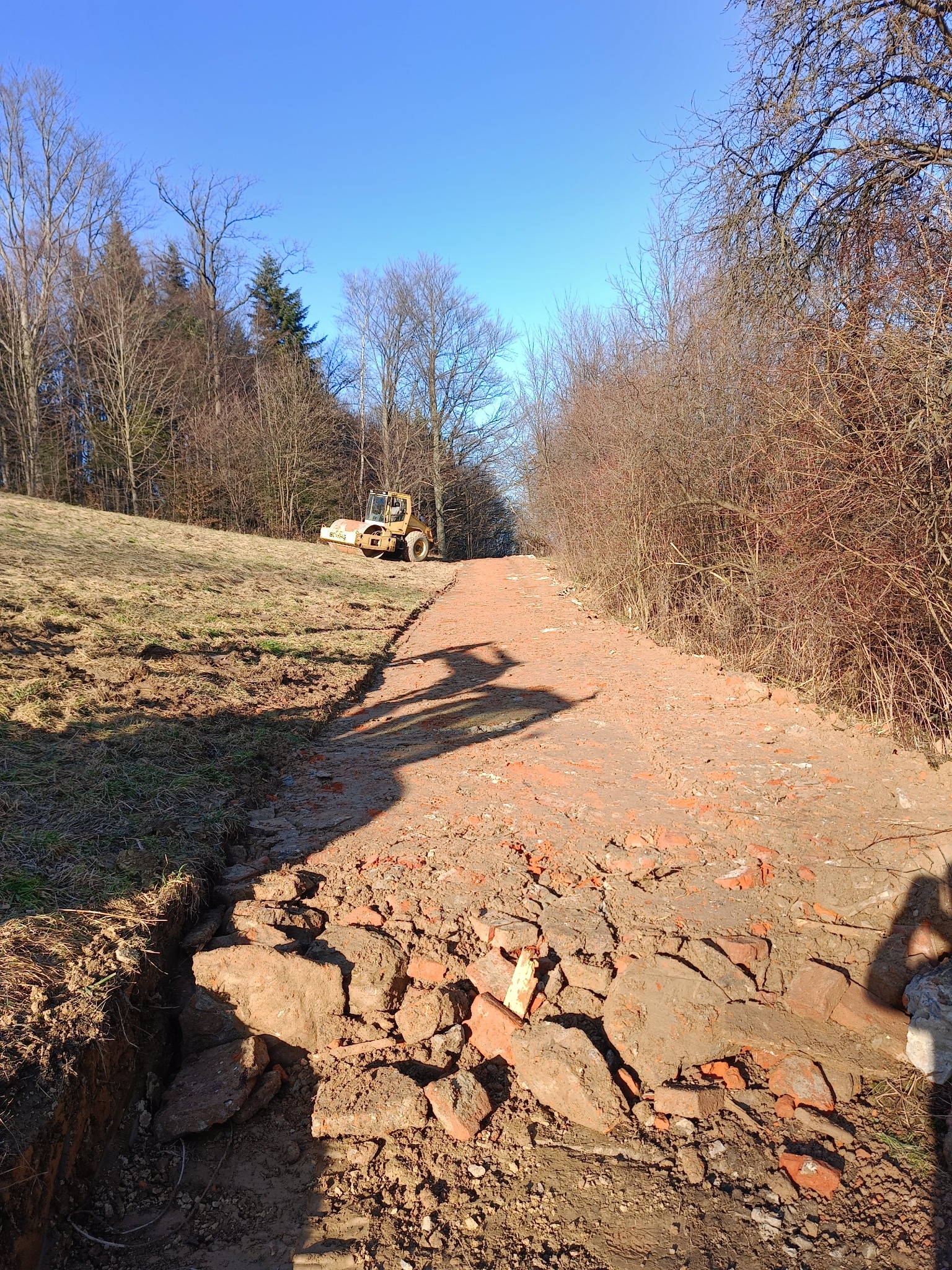 Żółty walec drogowy ugniata gruz ceglany na nierównej drodze gruntowej w słoneczny dzień, widoczny cień osoby w prawym dolnym rogu.