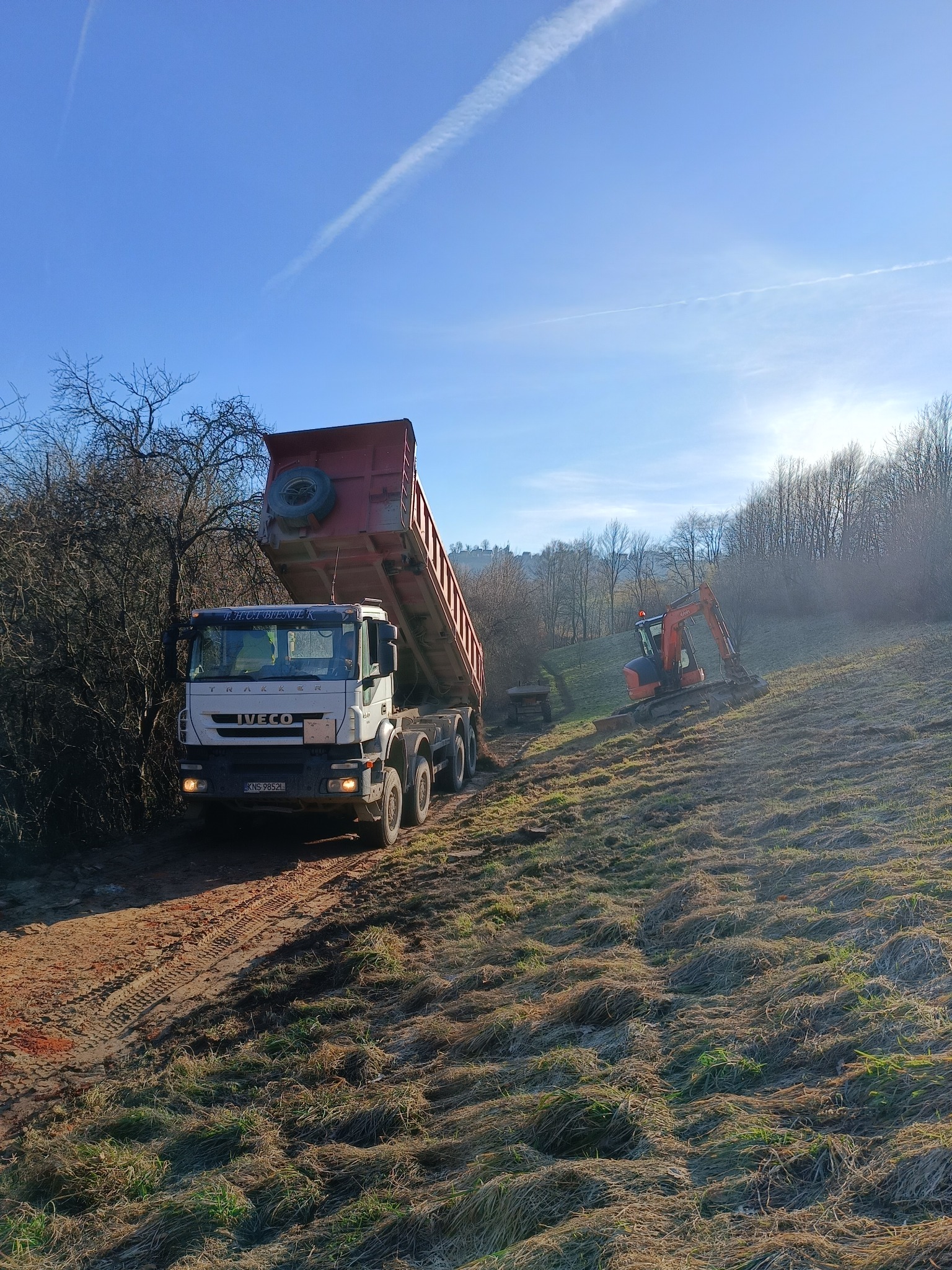 Ciężarówka z podniesioną skrzynią ładunkową i koparka pracują na zboczu w Nowym Sączu, pod błękitnym niebem z widocznymi smugami kondensacyjnymi.