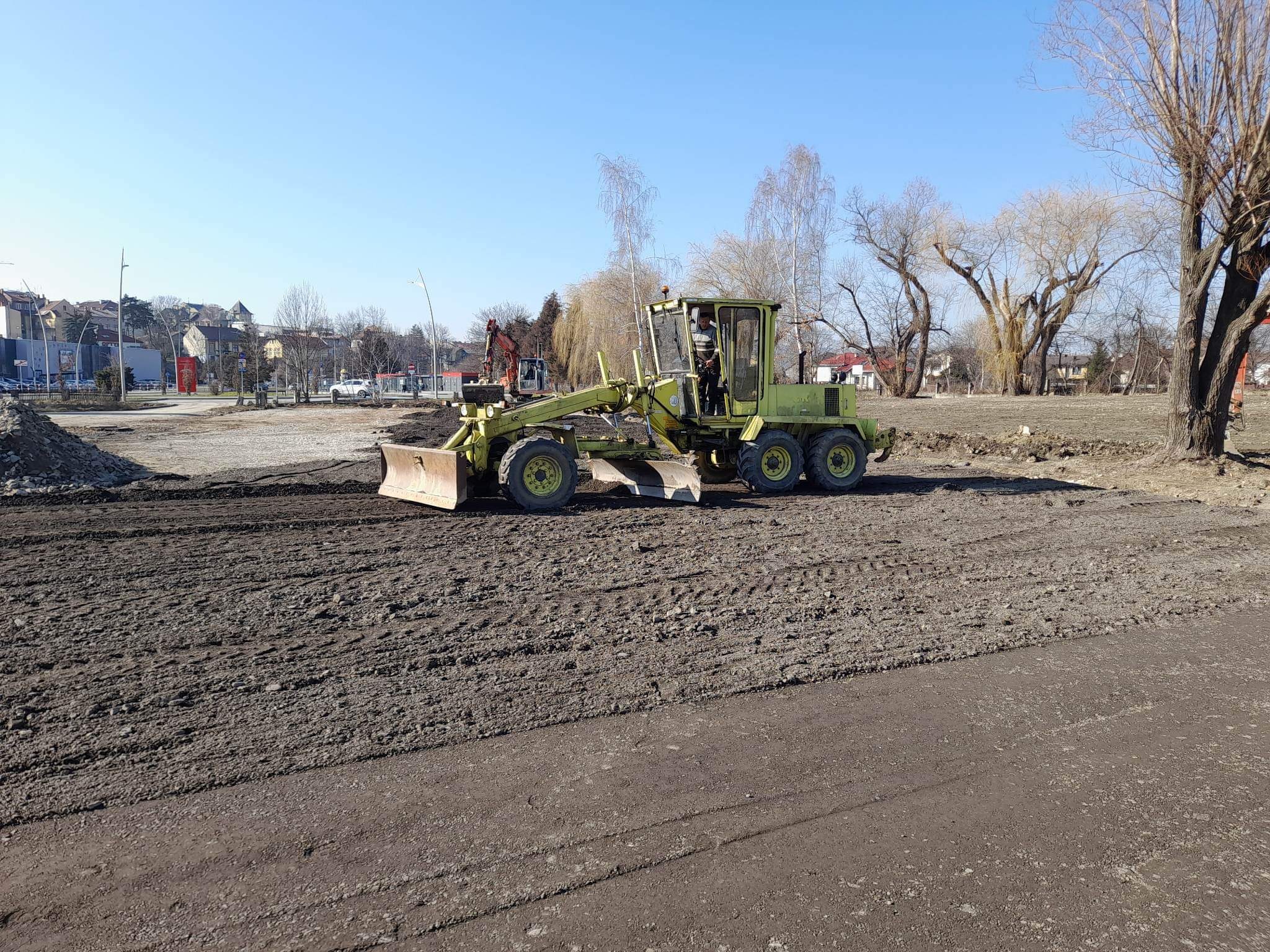 Żółty równiarka drogowa wyrównuje teren pod przyszłą drogę na placu budowy w słoneczny dzień. W tle widoczne drzewa i budynki miejskie.