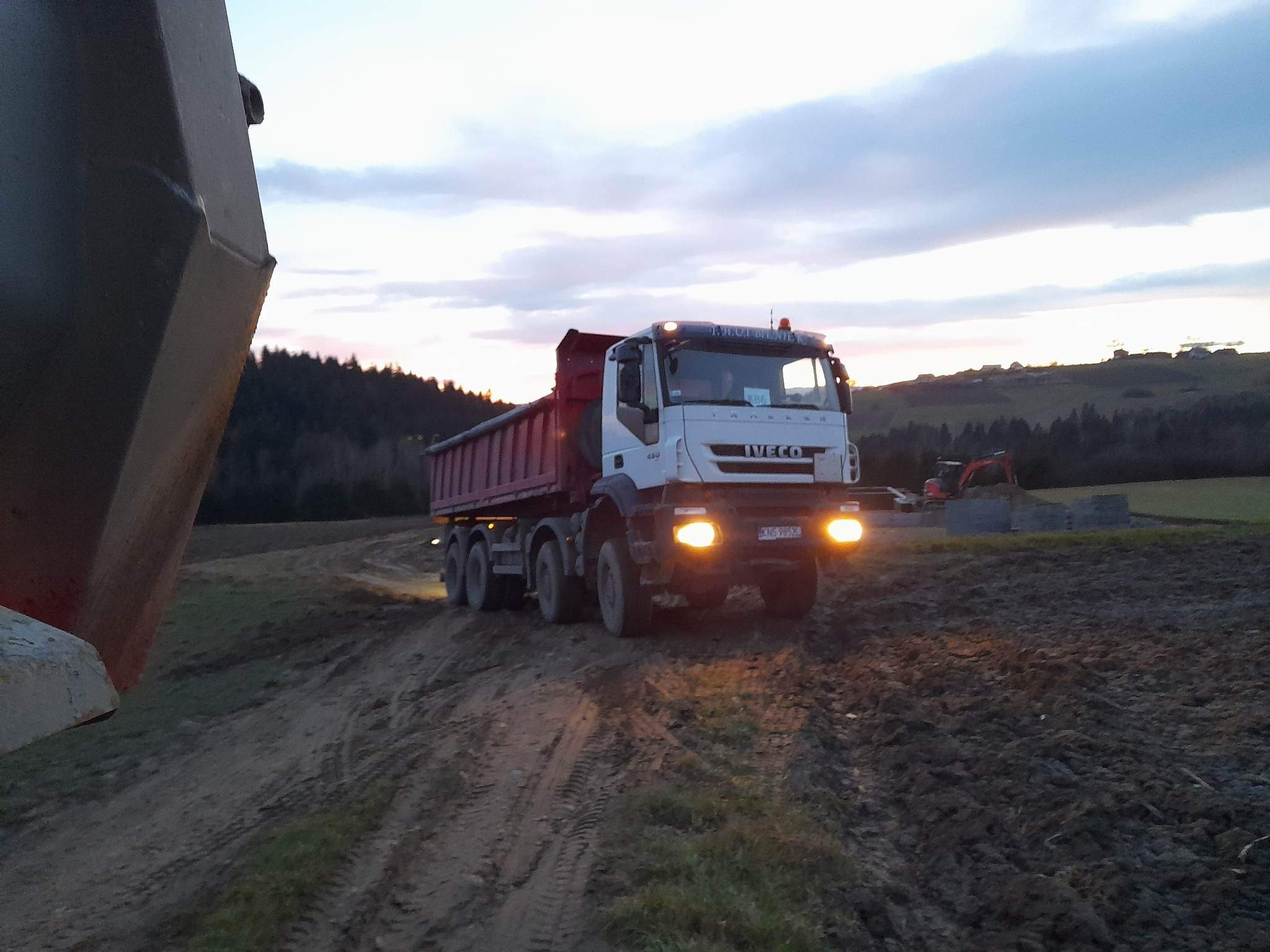 Ciężarówka Iveco z czerwoną skrzynią ładunkową na błotnistej drodze gruntowej o zmierzchu, w tle wzgórza i częściowo widoczna koparka.