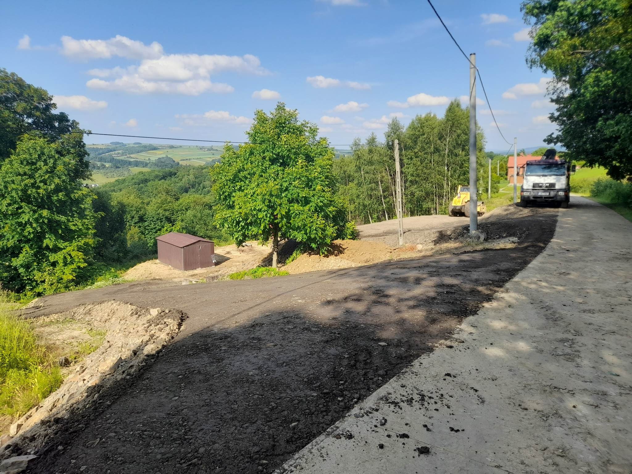 Walec drogowy i ciężarówka podczas prac na drodze gruntowej z widokiem na pagórkowaty krajobraz, mała brązowa budka w tle po lewej stronie.