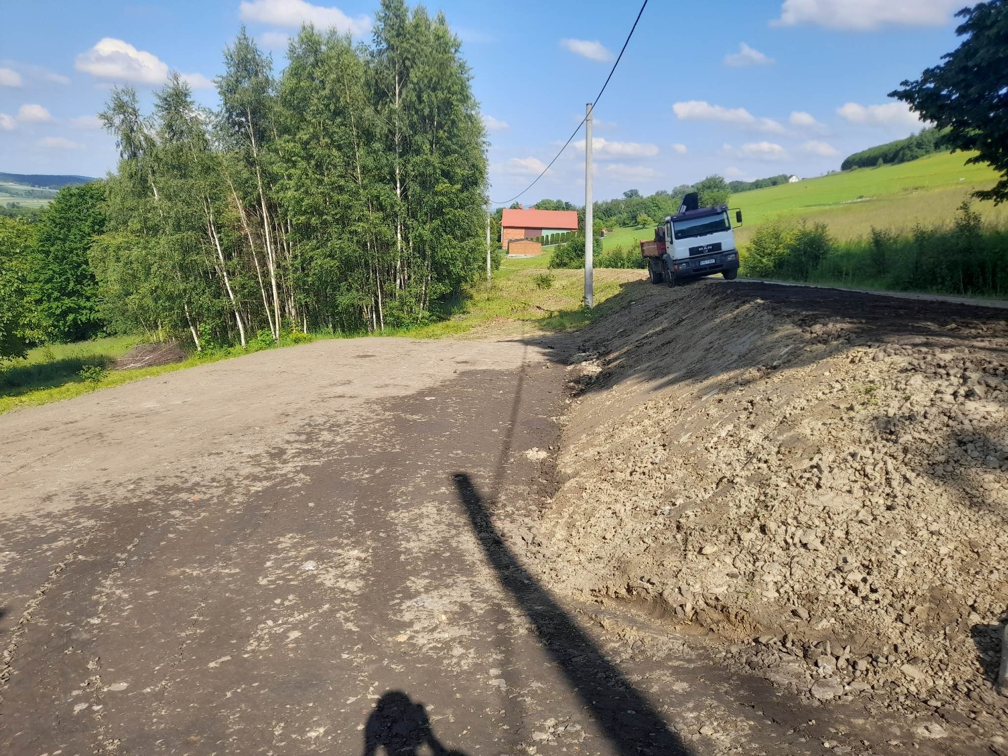 Ciężarówka MAN z wywrotką na drodze gruntowej w trakcie prac wyrównujących, widoczne świeże nasypy ziemi po bokach, w tle zieleń i zabudowania wiejskie, cień osoby w lewym dolnym rogu.