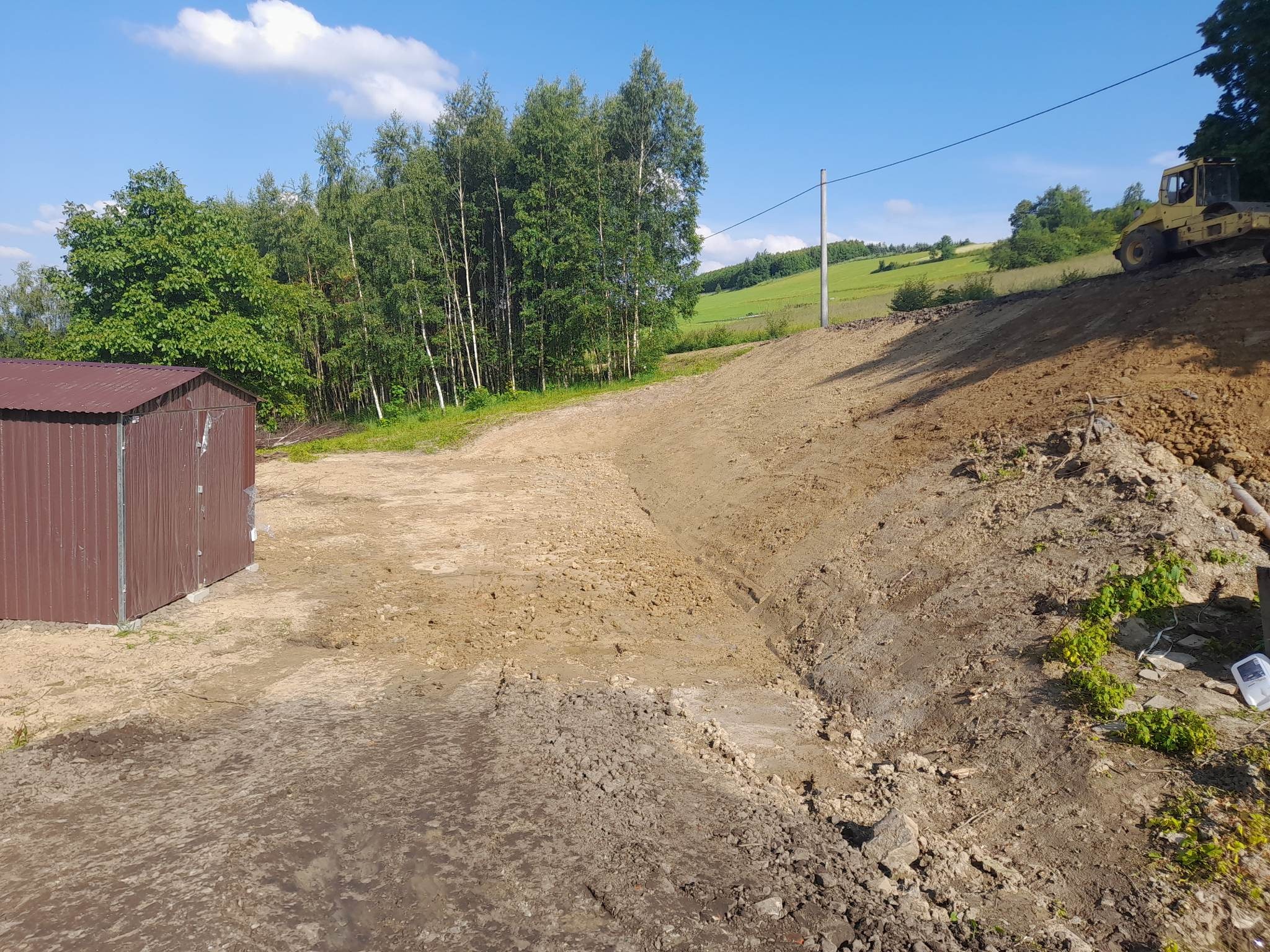 Żółty walec drogowy ubija ziemię na zboczu w słoneczny dzień, widoczny mały brązowy budynek gospodarczy, drzewa i zielone wzgórza w tle.