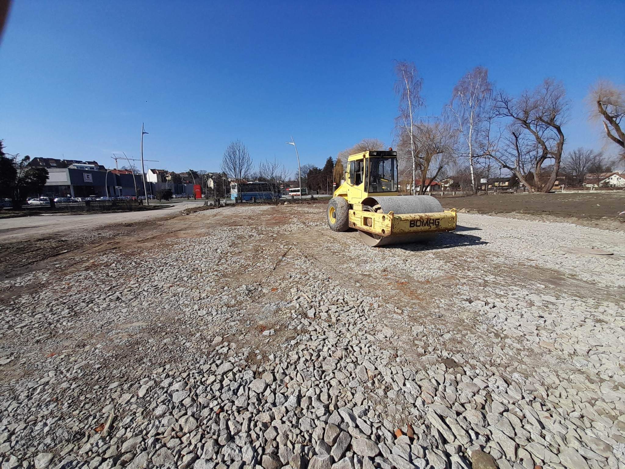 Żółty walec drogowy Bomag ugniata kamienistą nawierzchnię na placu budowy w słoneczny dzień, w tle budynki i drzewa.