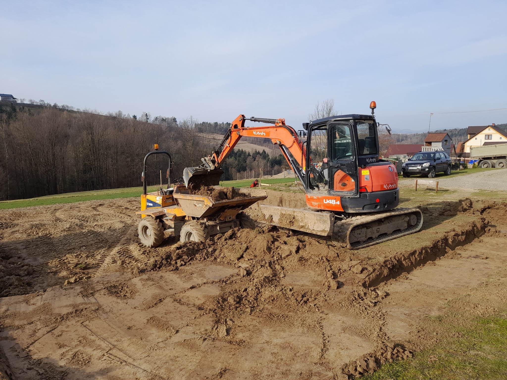 Pomarańczowa mini koparka Kubota U48-4 nabiera ziemię do żółtej wywrotki na placu budowy z widocznymi wykopami pod fundamenty, w tle drzewa i zabudowania.