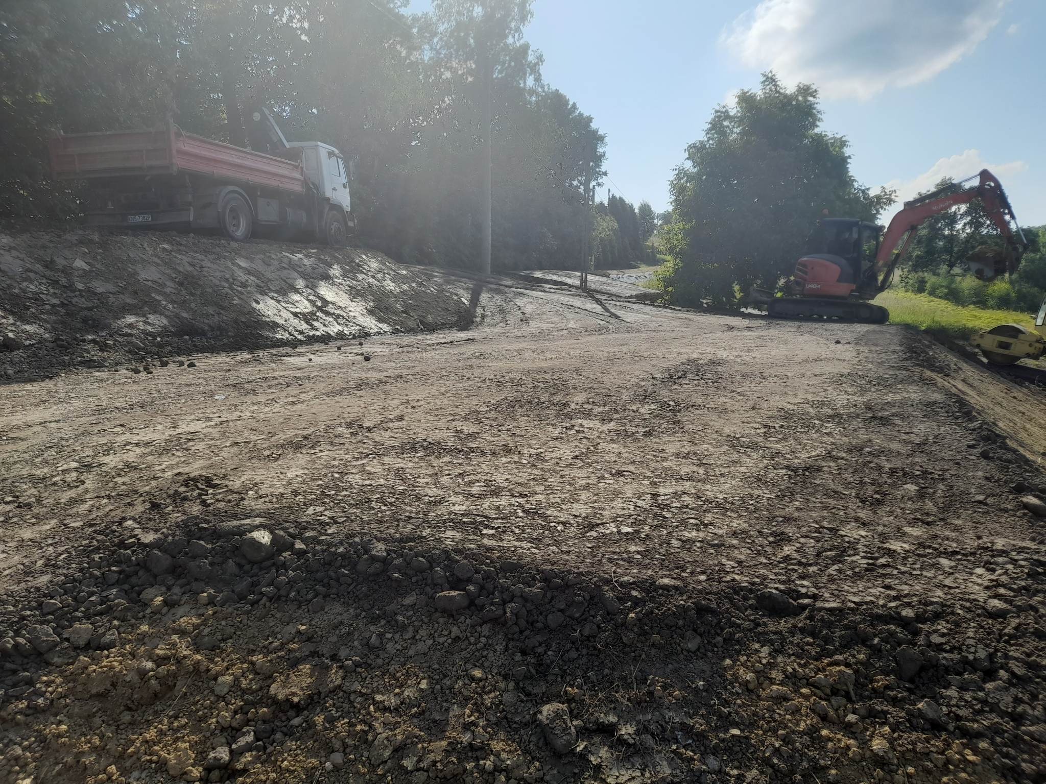 Przygotowanie terenu pod drogę: ciężarówka z czerwoną skrzynią na wzniesieniu, koparka Kubota U48-4 i walec drogowy na ubitej powierzchni, drzewa w tle.