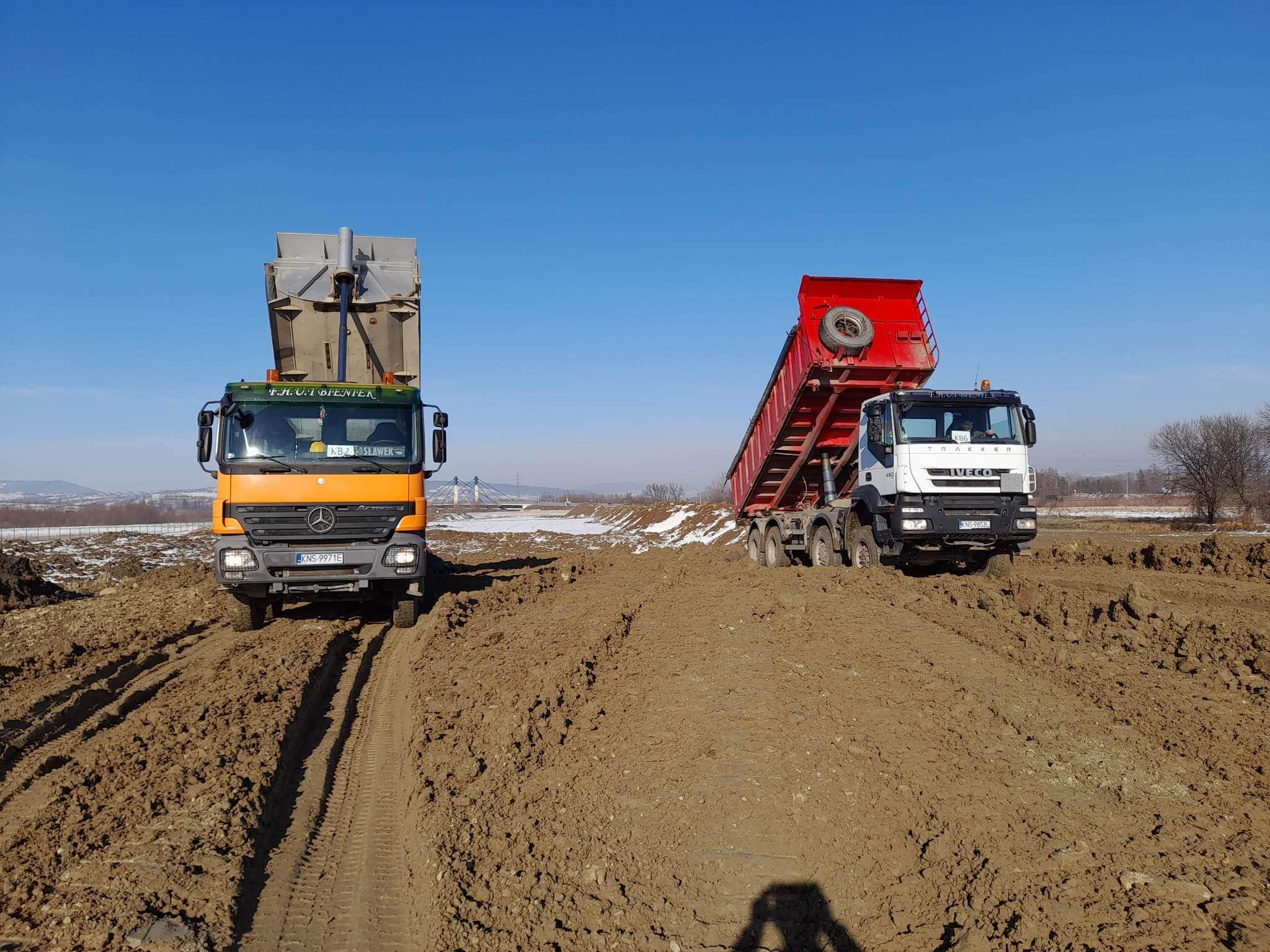 Dwa samochody ciężarowe (Mercedes i Iveco) stoją na nierównym, błotnistym terenie, prawdopodobnie placu budowy, pod błękitnym niebem; w tle widoczny most i częściowo ośnieżony krajobraz.