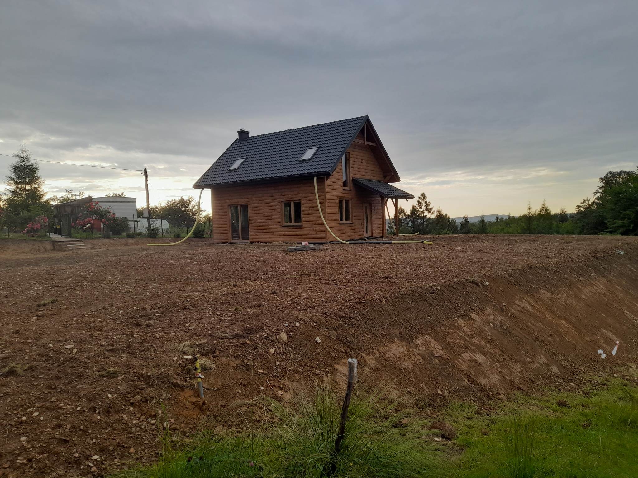 Drewniany dom z czarnym dachem na nierównym, przygotowanym terenie, z widocznymi żółtymi rurami, sugerującymi prace ziemne lub drenaż.