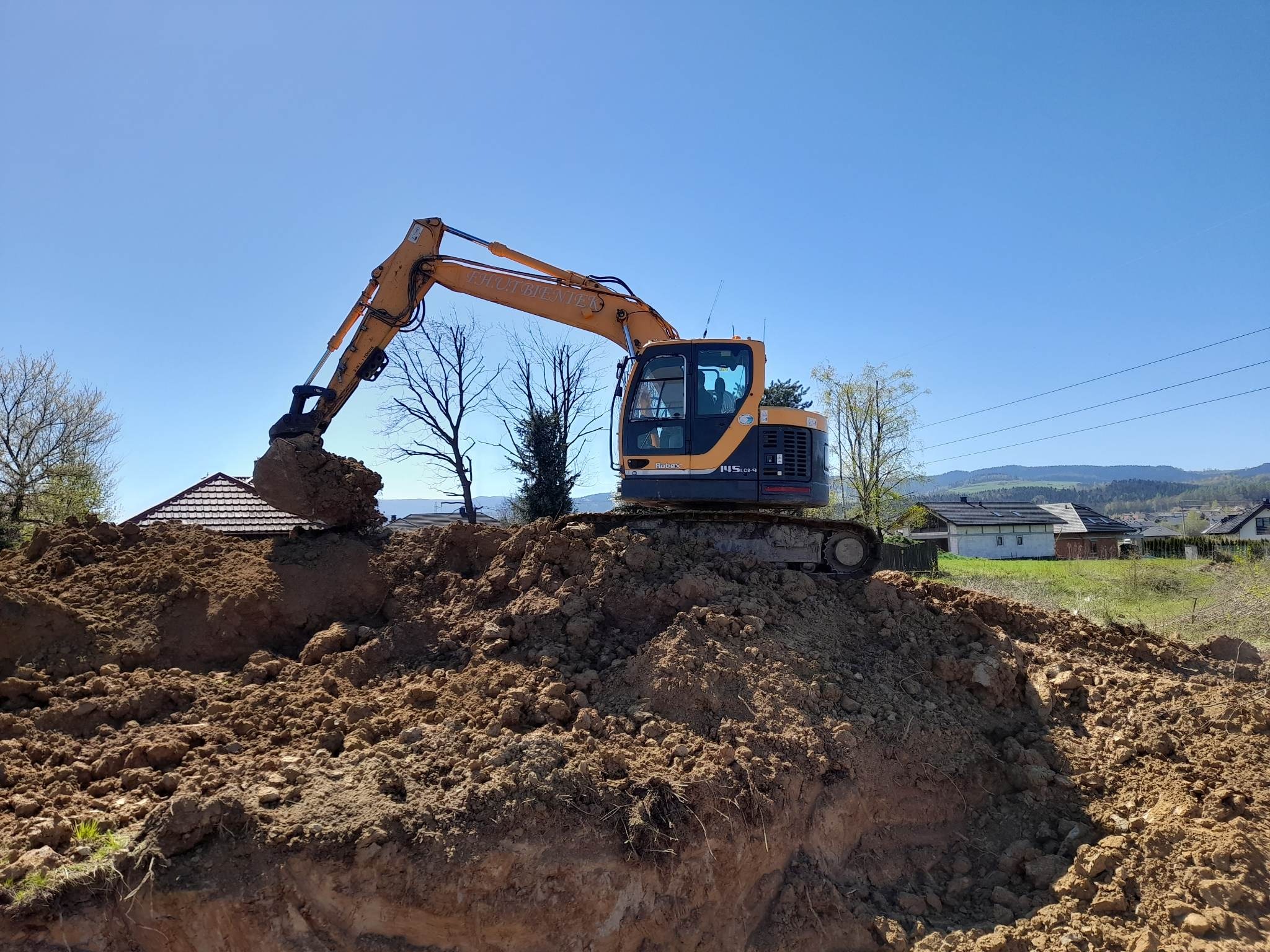 Żółta koparka Hyundai Robex 145CR-9 na wzniesieniu z ziemi, w tle domy i drzewa pod błękitnym niebem.