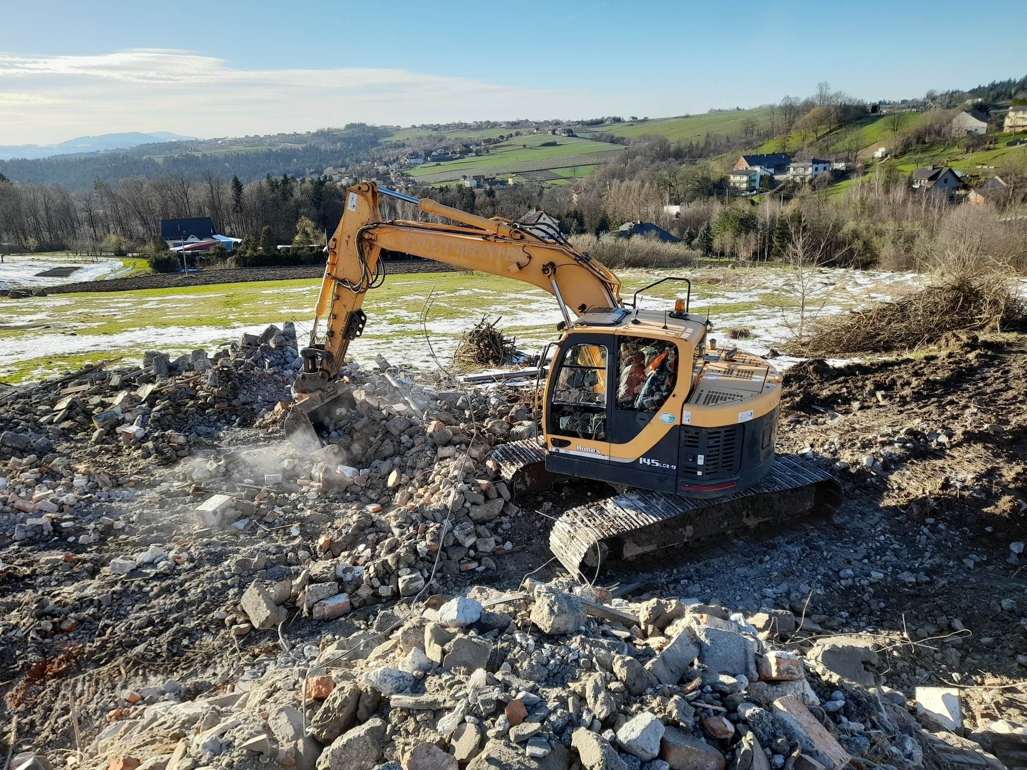 Żółta koparka Hyundai na gruzowisku po rozbiórce, w tle zielone wzgórza i fragmenty śniegu na łące.