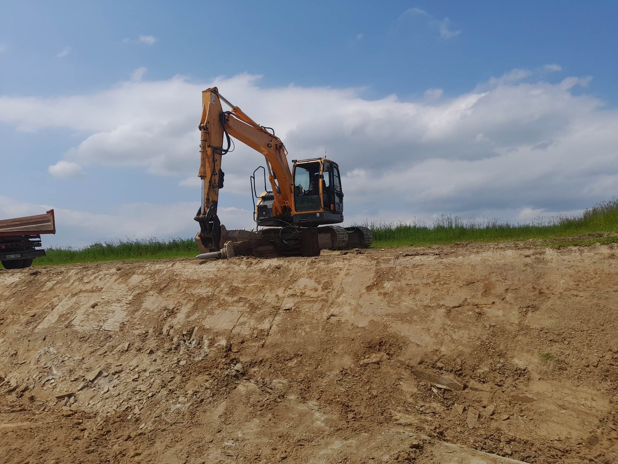 Żółta koparka hydrauliczna Hyundai z hydraulicznym młotem wyburzeniowym stoi na krawędzi wykopu, w tle zielona trawa i błękitne niebo z chmurami, z lewej strony fragment ciężarówki wywrotki.