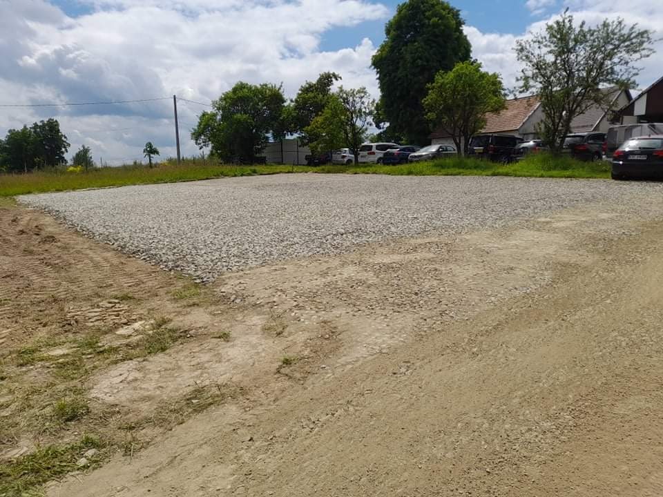 Utwardzony teren żwirowy pod parking, widoczne ślady opon, w tle zabudowania mieszkalne i zieleń, pochmurne niebo.