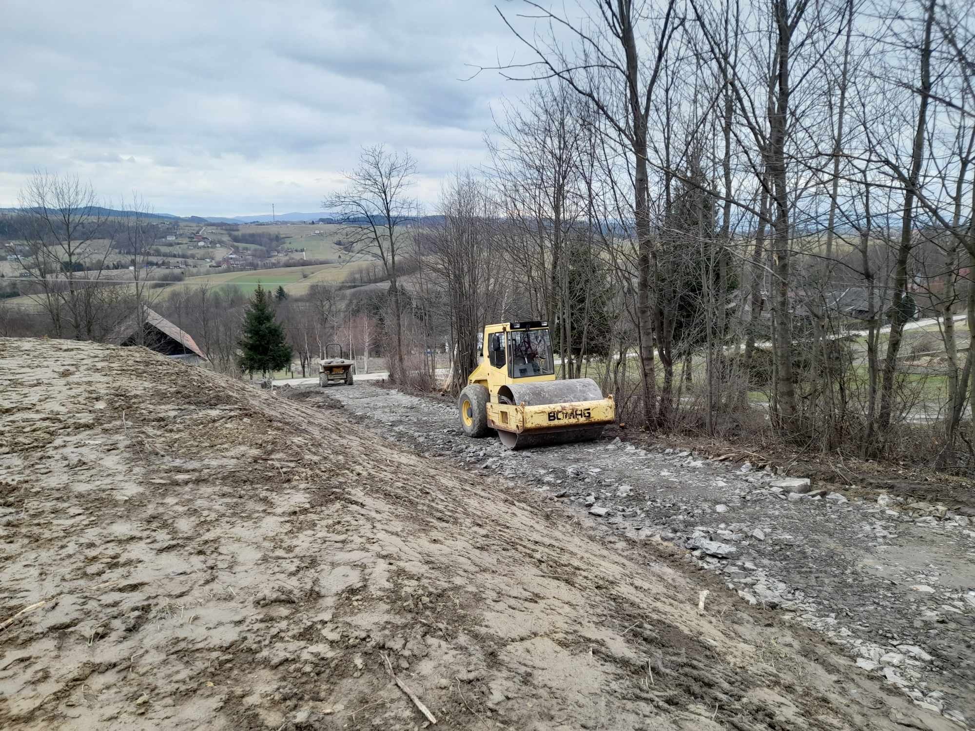 Żółty walec drogowy Bomag ubija kamienistą nawierzchnię na tle pagórkowatego krajobrazu z drzewami bez liści i zachmurzonym niebem.