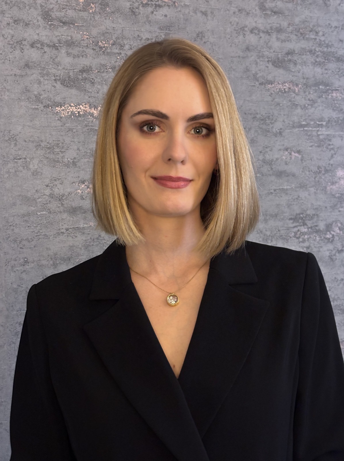 Portret kobiety w czarnej marynarce na tle szarej ściany. Kobieta ma blond włosy do ramion i delikatny makijaż. Uwagę przykuwa złoty naszyjnik z okrągłą zawieszką.