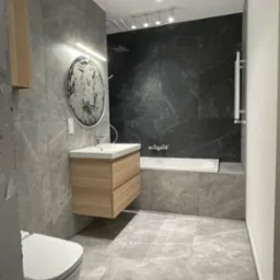 Nowoczesna łazienka w szarościach z drewnianą szafką pod umywalką, okrągłym lustrem z podświetleniem LED, czarną ścianą nad wanną i podwieszaną toaletą.