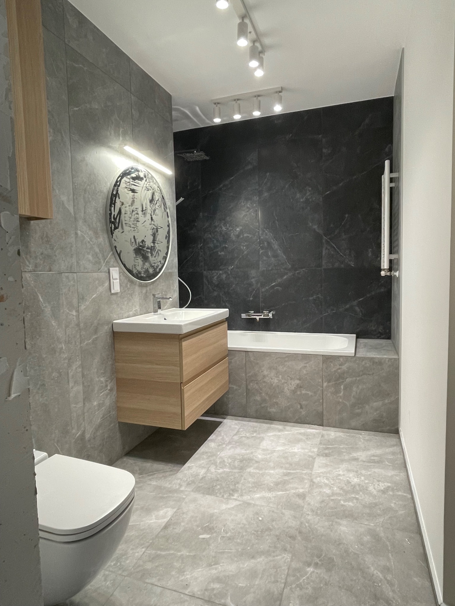 Nowoczesna łazienka w szarościach z drewnianą szafką pod umywalką, okrągłym lustrem z podświetleniem LED, czarną ścianą nad wanną i podwieszaną toaletą.