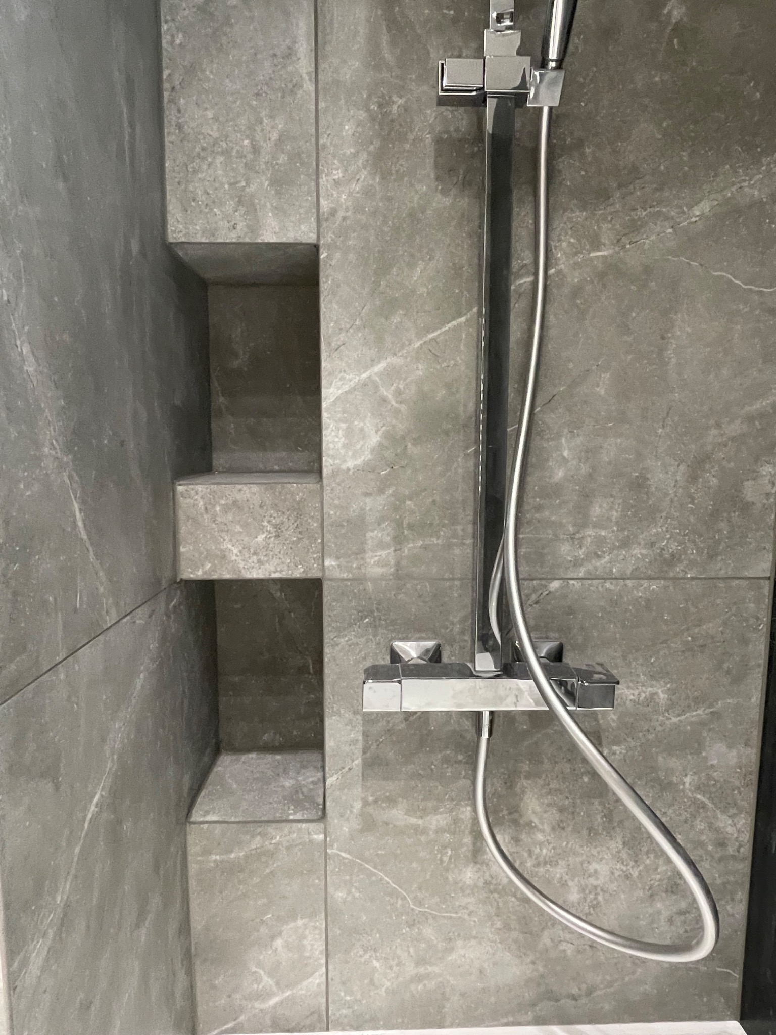 Nowoczesna kabina prysznicowa z szarymi płytkami imitującymi marmur i wnękami w ścianie, chromowana bateria natryskowa.