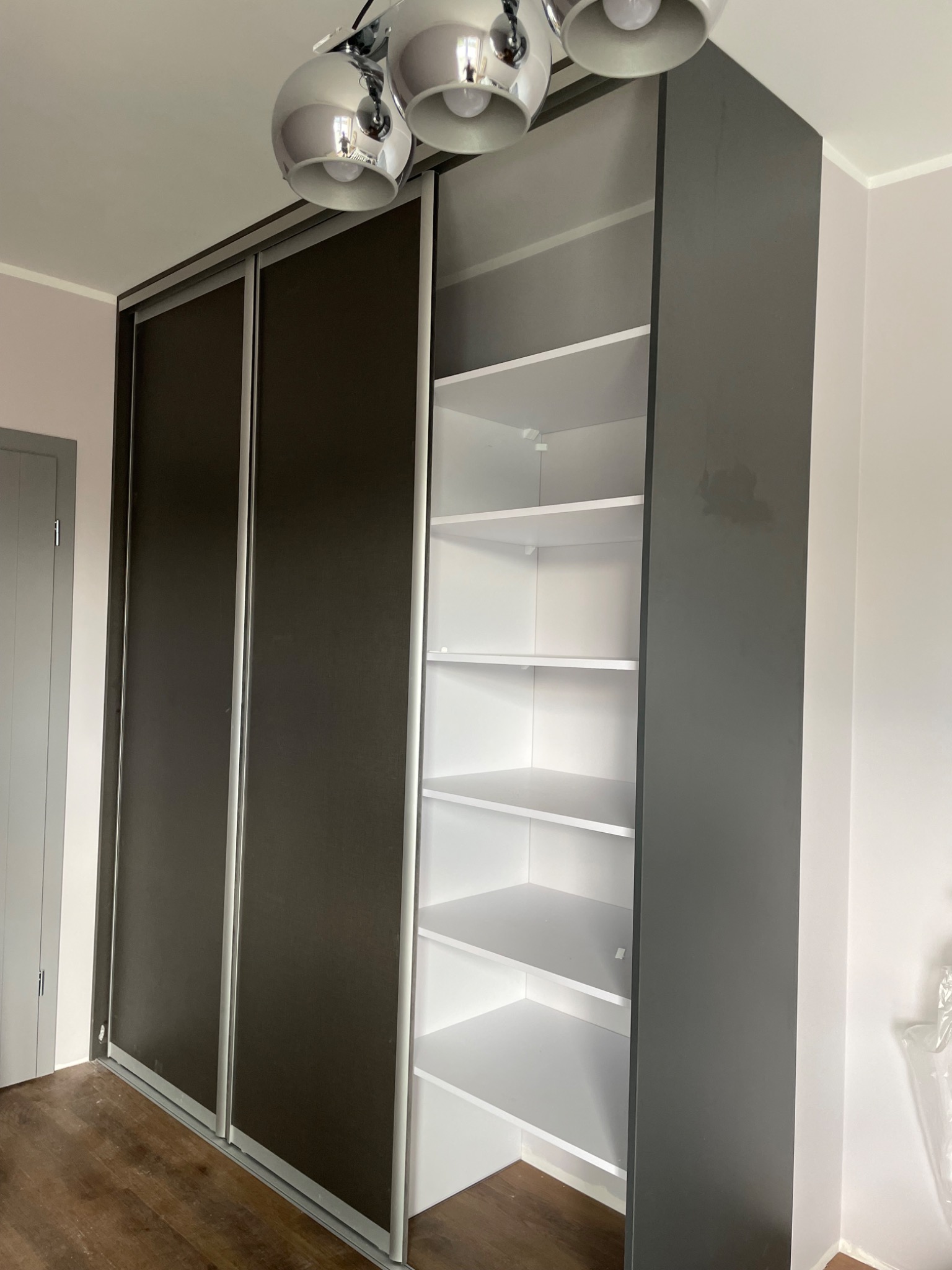 Wnętrze pokoju z dużą, nowoczesną szafą wnękową z przesuwnymi drzwiami w kolorze grafitowym i białymi półkami w otwartej części, oświetloną trzypunktową lampą sufitową w stylu glamour.