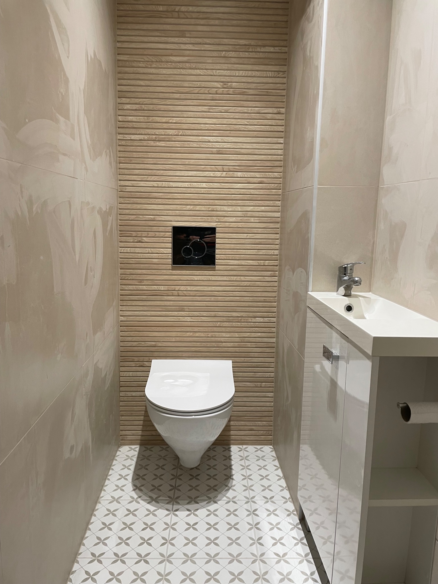 Nowoczesna, wykończona łazienka w Świętochłowicach z beżowymi płytkami imitującymi drewno na ścianie za toaletą, biała wisząca toaleta, biała szafka z umywalką i wzorzysta podłoga.