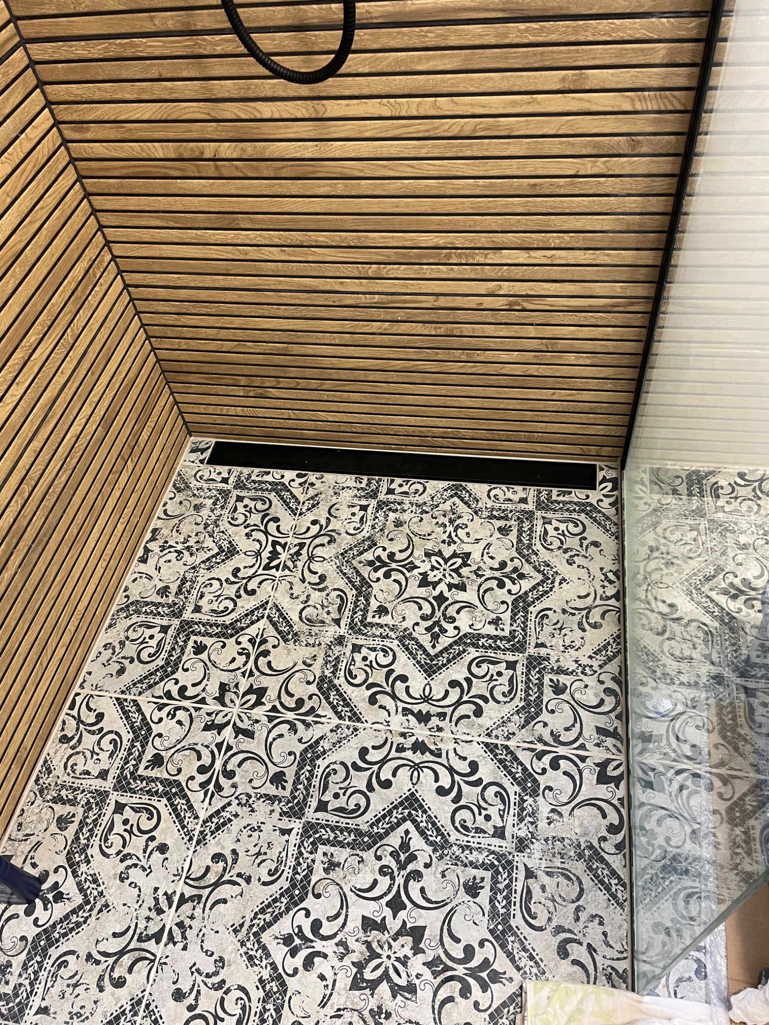 Nowoczesna łazienka z drewnianą boazerią na ścianach i suficie oraz wzorzystą, czarno-białą podłogą i szklaną kabiną prysznicową.