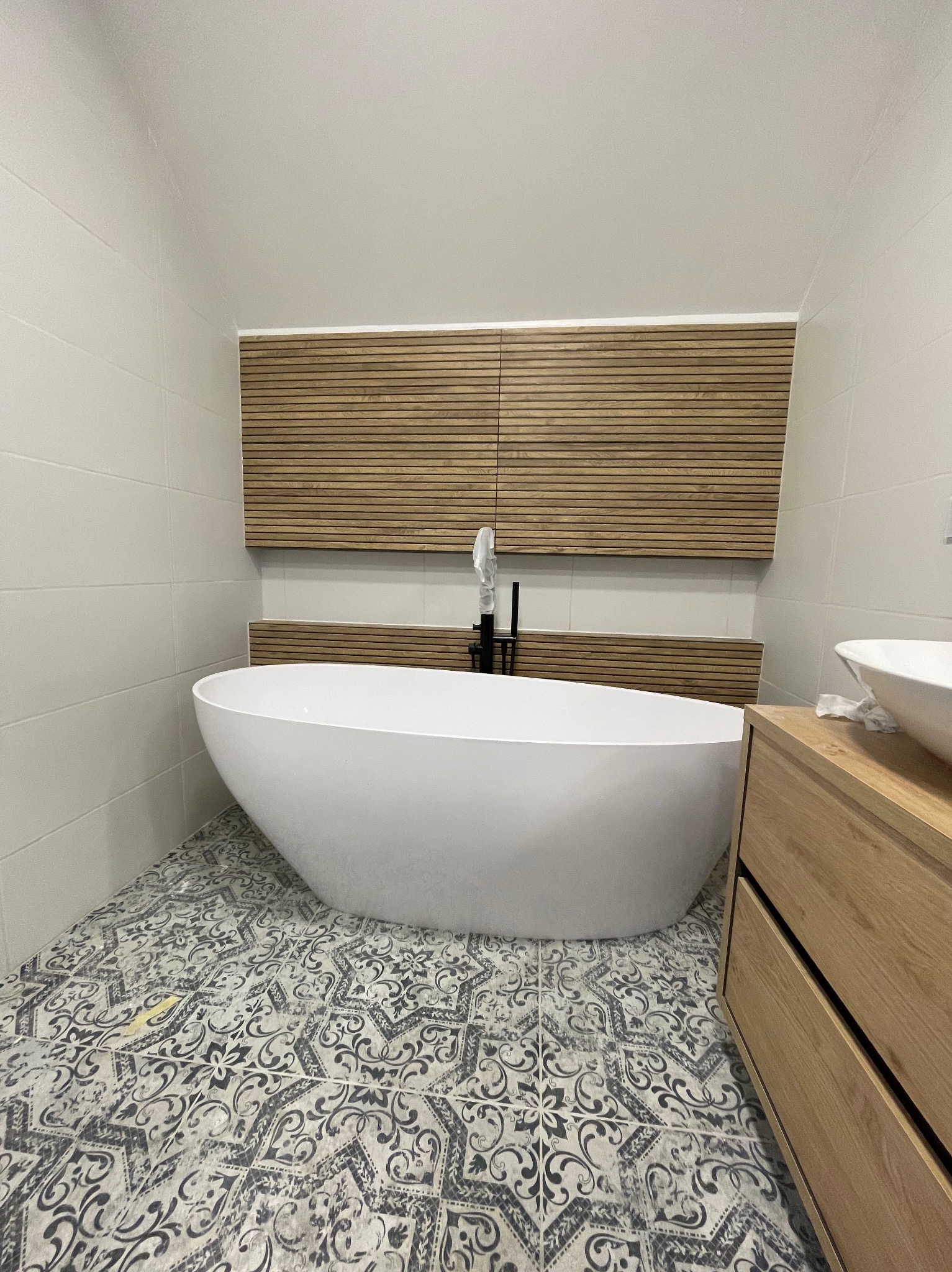 Nowoczesna łazienka z wolnostojącą białą wanną, czarną baterią umywalkową, drewnianą szafką pod umywalką i dekoracyjnym panelem ściennym z drewnianych listew, na podłodze wzorzyste płytki.