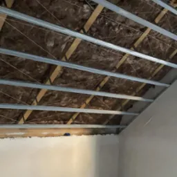 Poddasze z widoczną izolacją z wełny mineralnej, metalowym stelażem i drewnianymi belkami, przygotowane do montażu płyt kartonowo-gipsowych. Widoczny fragment ściany.