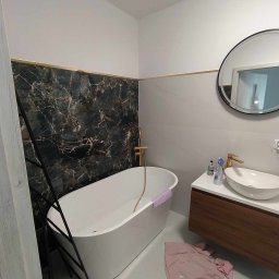 Ka-bud - Nowoczesna łazienka z czarnym marmurem i złotymi akcentami, biała wanna wolnostojąca z złotą baterią, okrągłe lustro nad umywalką, różowy ręcznik na podłodze.