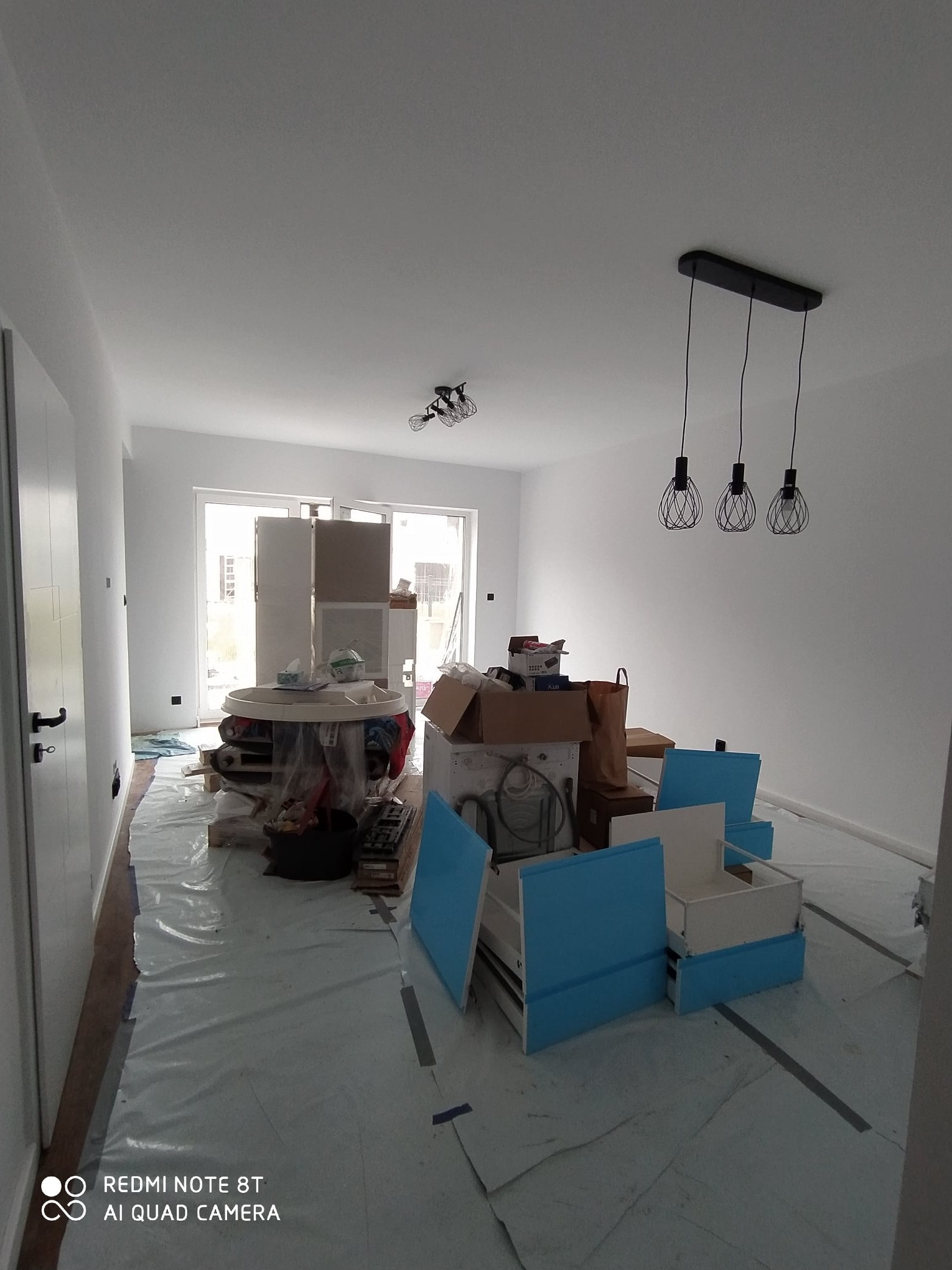 Wnętrze w trakcie prac remontowych, zabezpieczona podłoga folią, elementy mebli kuchennych w kolorze błękitnym, kartony, okrągły stół i nowoczesne lampy wiszące.