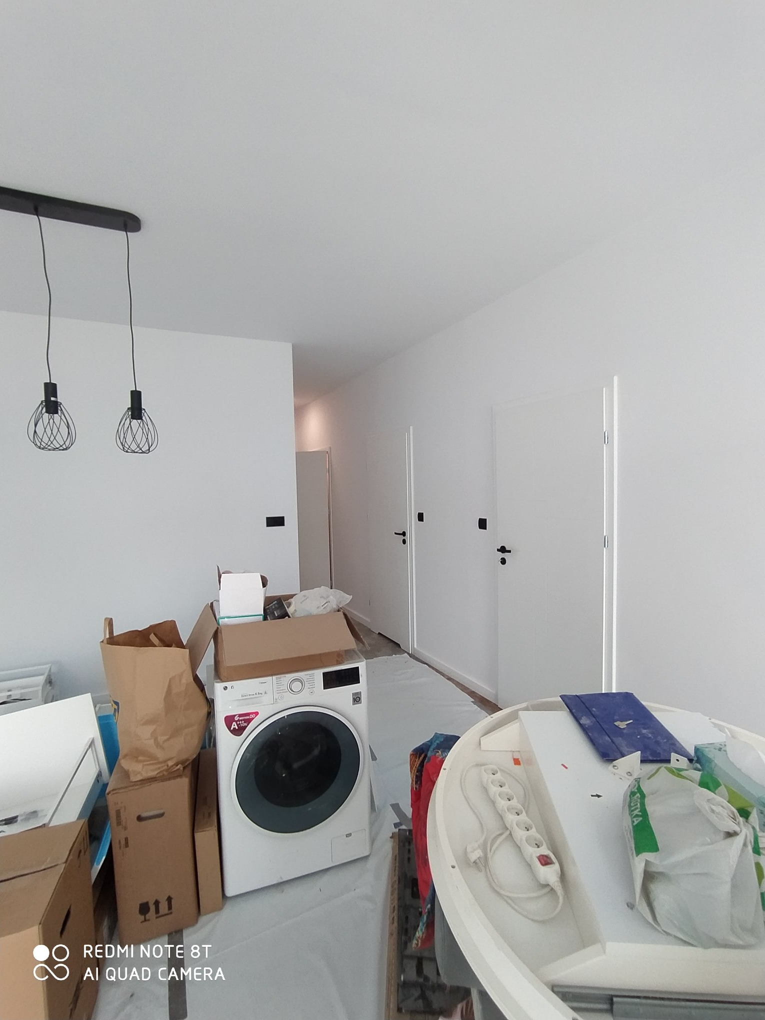 Wnętrze w trakcie prac wykończeniowych: białe ściany, wiszące lampy, nowa pralka otoczona kartonami, zabezpieczona podłoga, widoczne narzędzia i materiały budowlane.