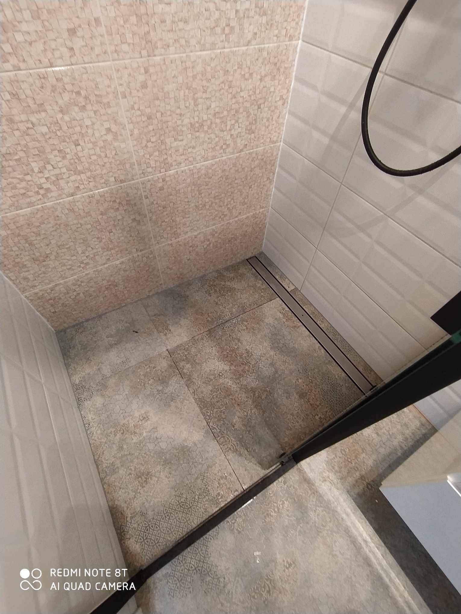 Wykończony prysznic z mozaiką na górnej części ściany, dużymi białymi płytkami na dolnej części ściany i wzorzystymi płytkami podłogowymi oraz liniowym odpływem.