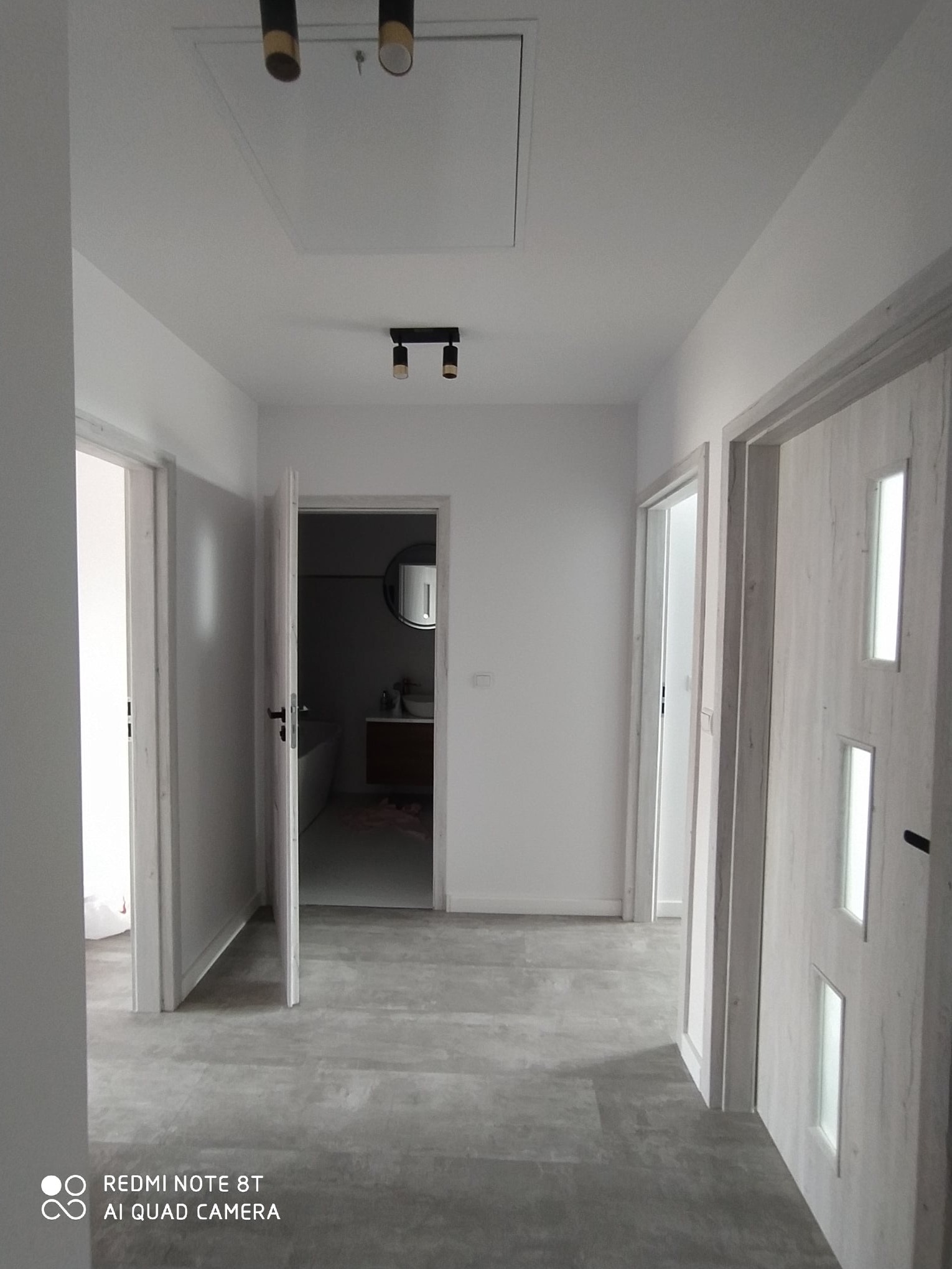 Korytarz w jasnych barwach z otwartymi drzwiami do łazienki, widoczne szare płytki podłogowe imitujące drewno, białe ściany i drzwi z przeszkleniami.