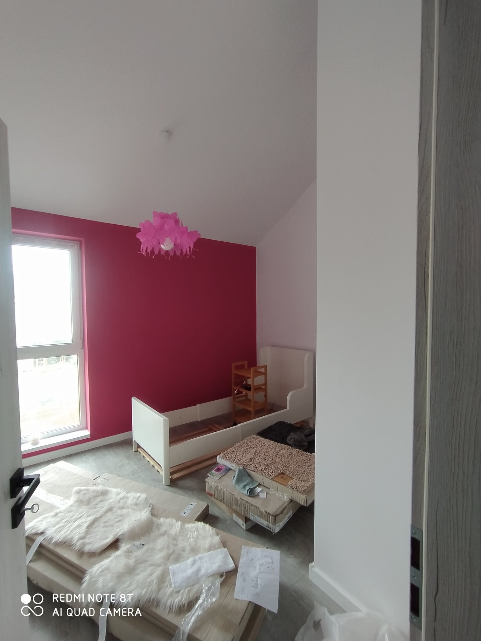 Pokój dziecięcy w trakcie urządzania: ściana pomalowana na intensywny róż, białe łóżko w trakcie montażu, różowy abażur, dywaniki i kartony na podłodze, widoczny fragment drzwi z szarą okleiną.