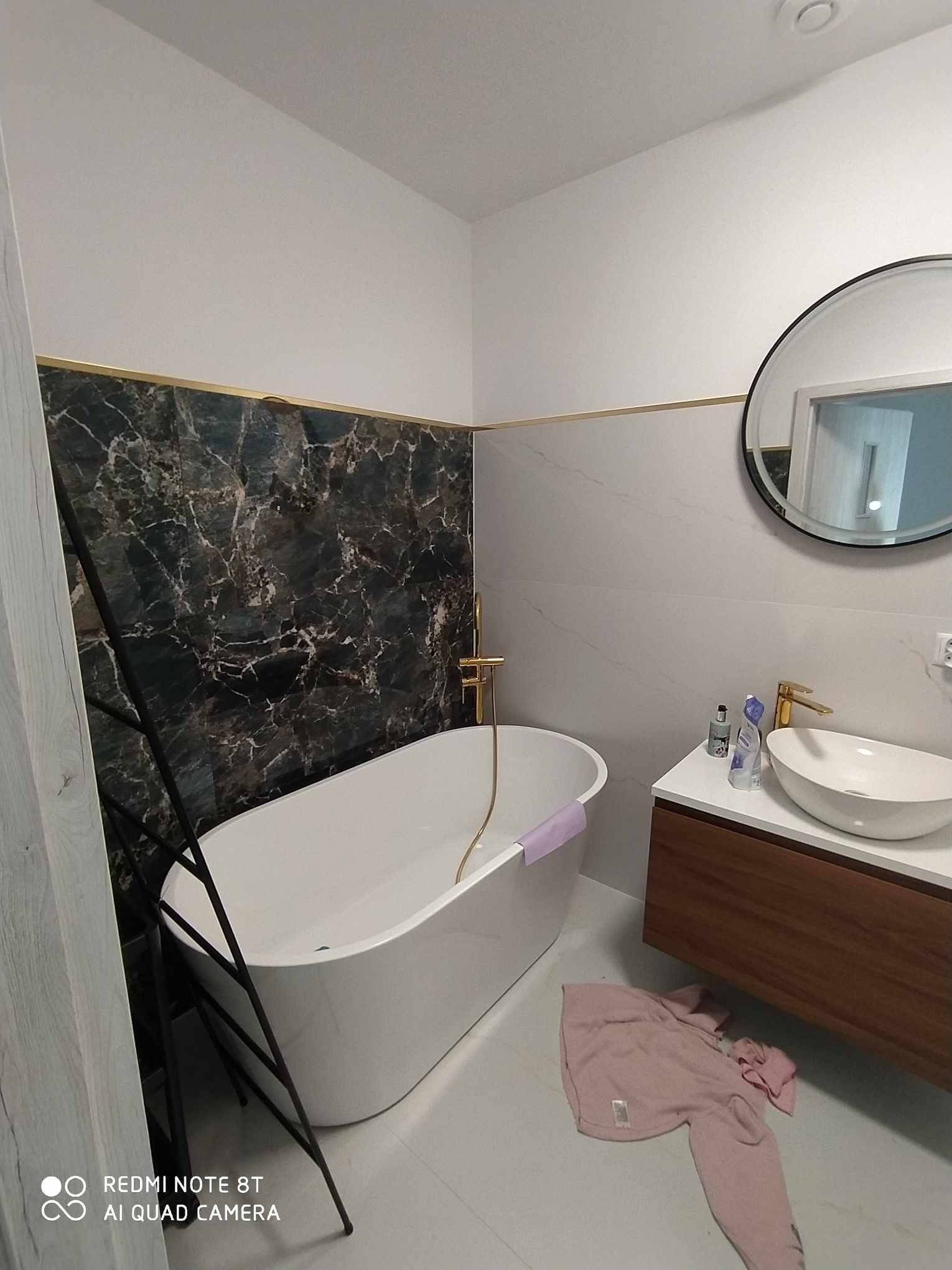 Nowoczesna łazienka z czarnym marmurem i złotymi akcentami, biała wanna wolnostojąca z złotą baterią, okrągłe lustro nad umywalką, różowy ręcznik na podłodze.