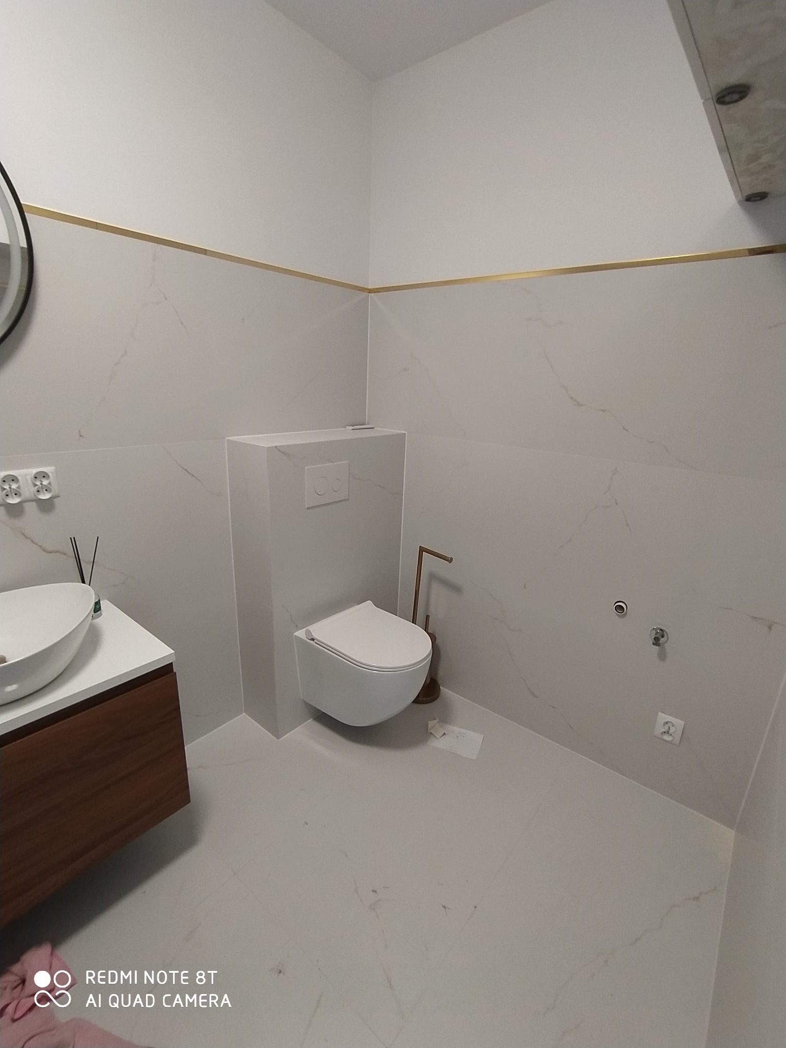 Nowoczesna łazienka z białymi płytkami imitującymi marmur, złotym akcentem w postaci listwy, podwieszanym WC i umywalką nablatową na drewnianej szafce.
