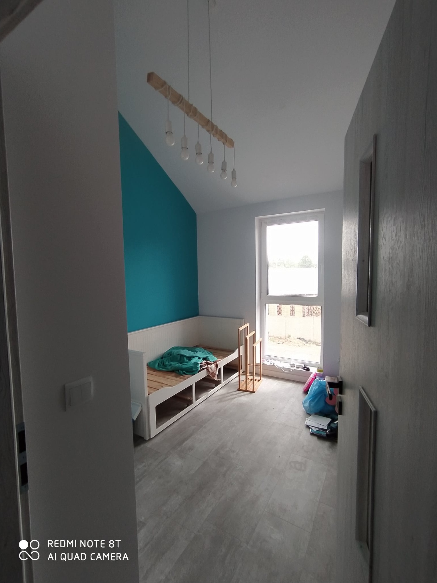 Pokój po odświeżeniu z turkusową ścianą, drewnianym łóżkiem z szufladami i wiszącą lampą z żarówkami na sznurkach, widoczne ślady po remoncie.