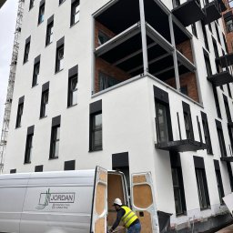 Jordan Construction & Consulting - Pracownik w kamizelce odblaskowej wyjmuje materiały z furgonetki z logo firmy na tle nowego budynku wielorodzinnego z balkonami i oknami w czarnych obramowaniach.