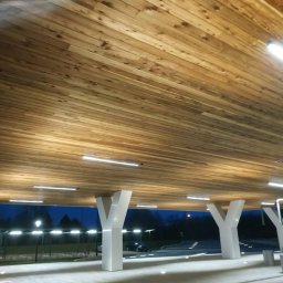 Jordan Construction & Consulting - Drewniany sufit podwieszany z oświetleniem LED w przestrzeni zewnętrznej z białymi kolumnami w kształcie litery Y, widok na zewnątrz o zmierzchu.