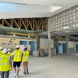 Jordan Construction & Consulting - Grupa inspektorów w kamizelkach odblaskowych i kaskach ogląda stan surowy biura przed remontem, widoczne odsłonięte elementy konstrukcyjne stropu, ściany z surowego betonu komórkowego oraz worki...