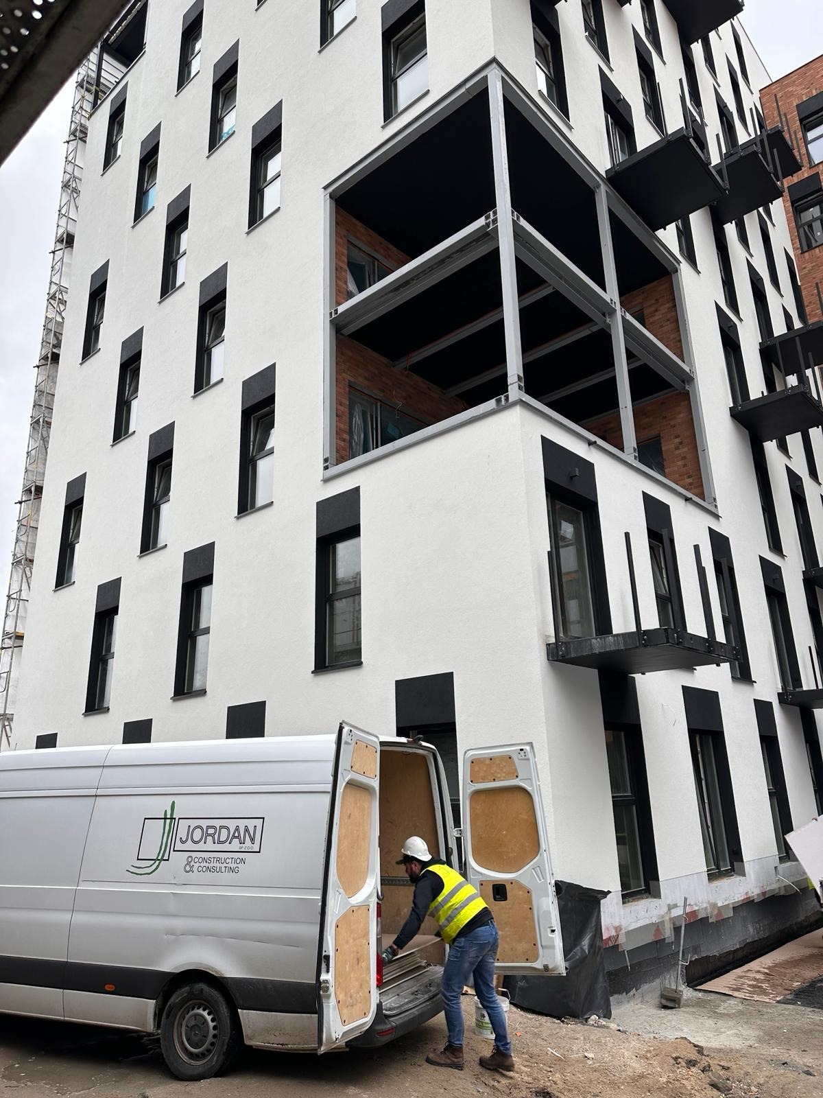 Pracownik w kamizelce odblaskowej wyjmuje materiały z furgonetki z logo firmy na tle nowego budynku wielorodzinnego z balkonami i oknami w czarnych obramowaniach.
