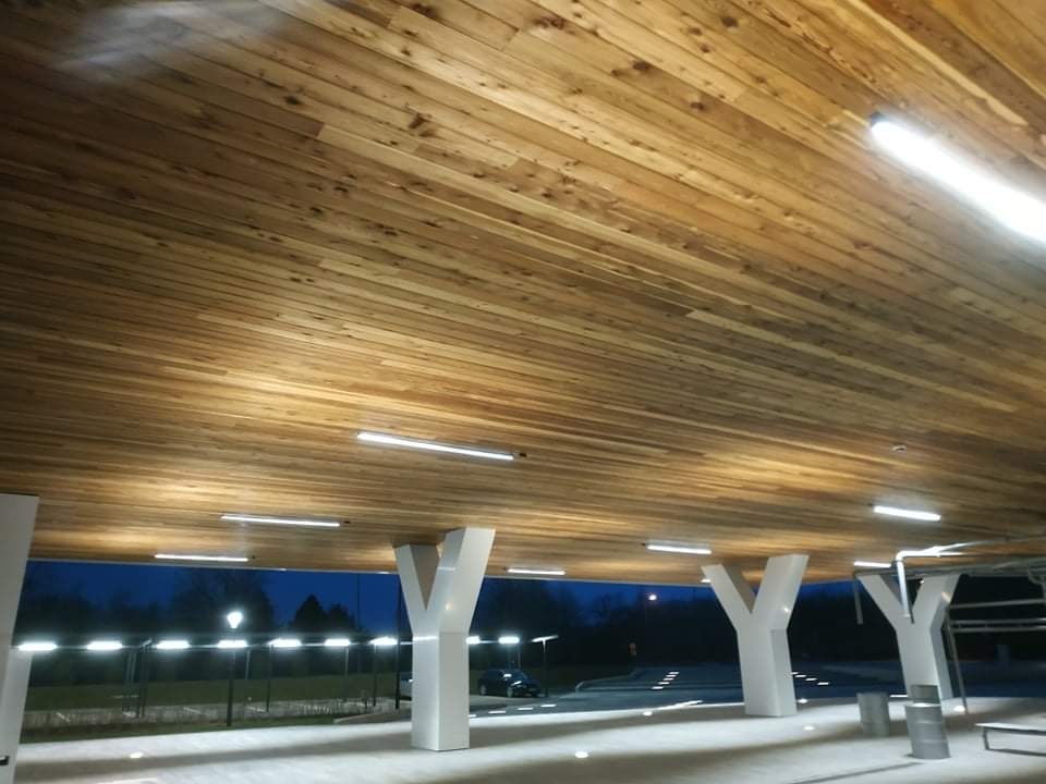 Drewniany sufit podwieszany z oświetleniem LED w przestrzeni zewnętrznej z białymi kolumnami w kształcie litery Y, widok na zewnątrz o zmierzchu.