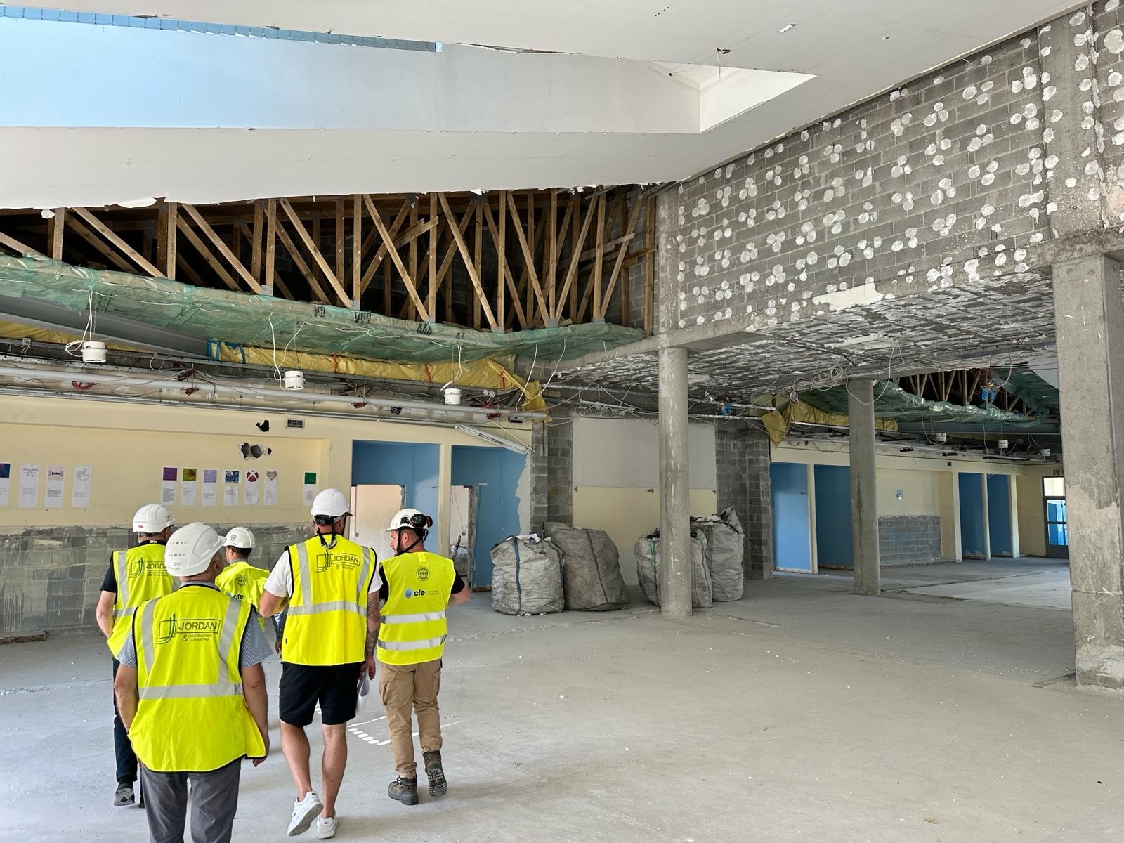 Grupa inspektorów w kamizelkach odblaskowych i kaskach ogląda stan surowy biura przed remontem, widoczne odsłonięte elementy konstrukcyjne stropu, ściany z surowego betonu komórkowego oraz worki...