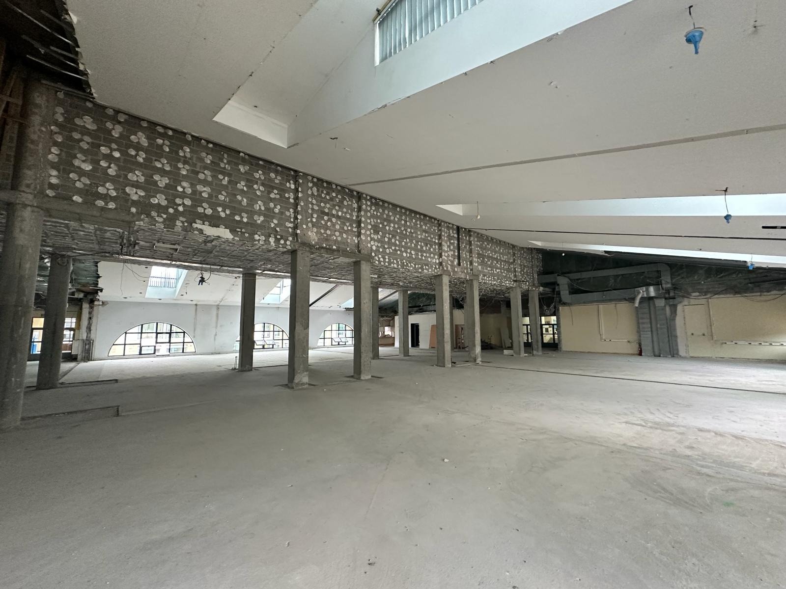 Wnętrze dużego, pustego biura w trakcie remontu. Widoczne betonowe kolumny podtrzymujące strop, odsłonięte instalacje i surowe ściany. Okna w kształcie łuków w oddali wpuszczają światło...