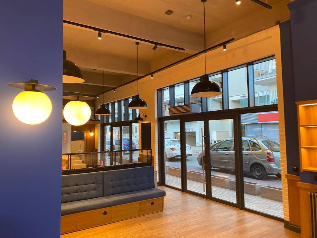 Wnętrze biura z drewnianą podłogą, niebieską tapicerowaną ławką, przeszkloną ścianą z widokiem na ulicę i nowoczesnymi, kulistymi lampami.