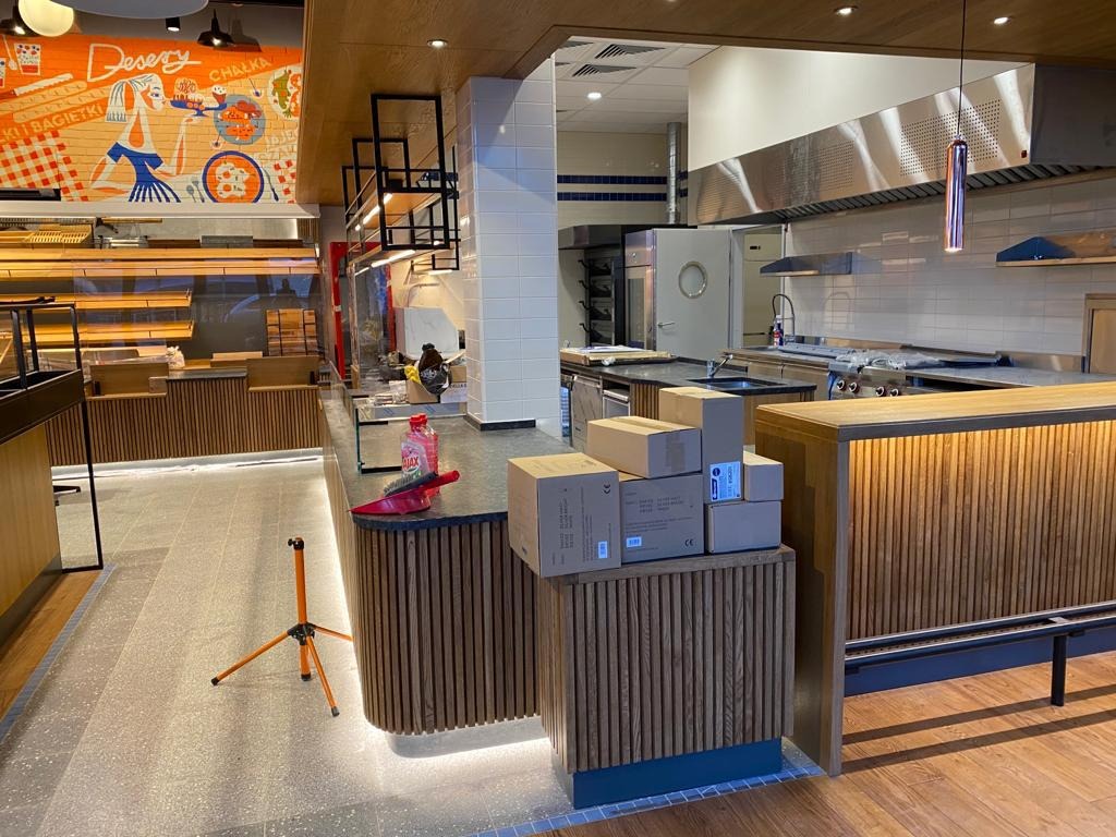 Wnętrze lokalu gastronomicznego w trakcie wykańczania: widoczna lada z drewnianą okładziną, kartony z produktami, sprzęt do sprzątania, w tle zaplecze kuchenne ze stalowymi urządzeniami i fragment...