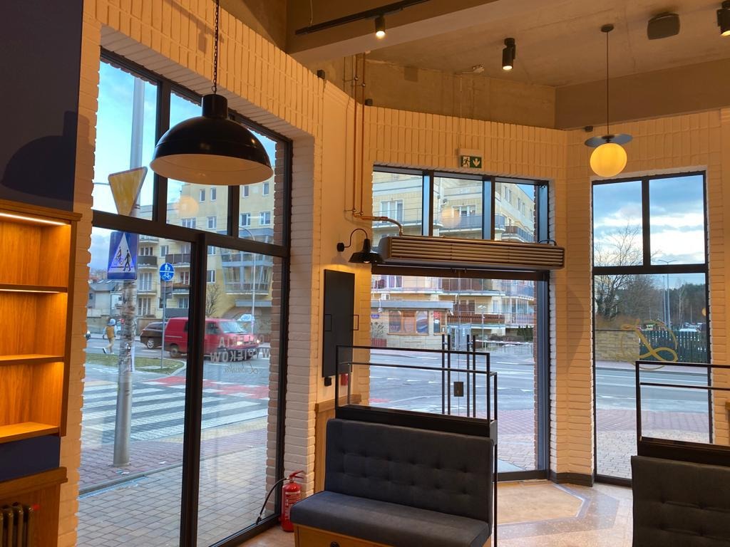 Wnętrze lokalu z widocznymi elementami wykończenia: białe cegły na ścianach, duże okna z czarnymi ramami, wiszące lampy i ciemnoszare kanapy. Na zewnątrz widoczny ruch uliczny i budynki.