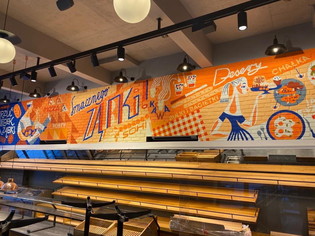 Wnętrze piekarni z pustymi półkami na pieczywo i muralem na ścianie z cegły, przedstawiającym motywy piekarnicze w pomarańczowo-niebieskiej kolorystyce.